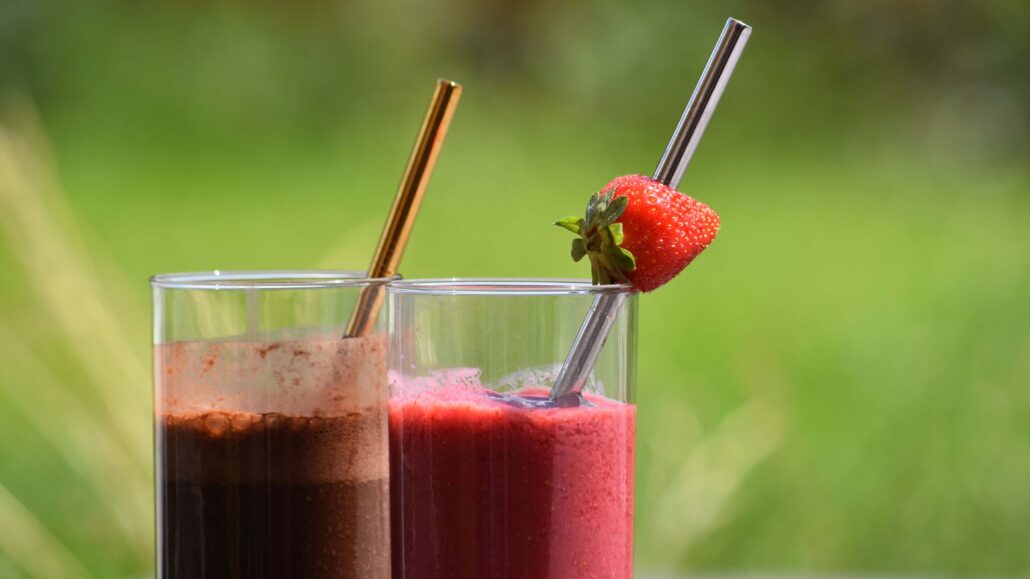 Image of a vegan strawberry milkshake with silver straw and a strawberry, and a vegan mokka milkshake with a golden straw, against a green outdoor background
