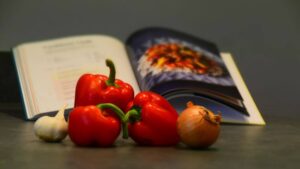 Image of three red peppers, an onion and garlic, in front of a Fork Ranger Book, opened to the page containing a recipe for Caribbean Chilli.