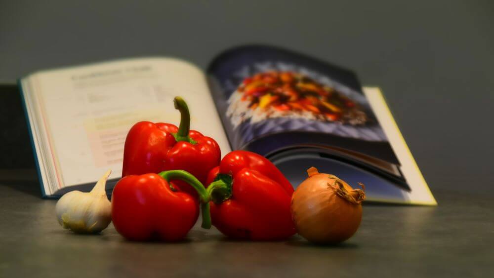 Image of three red peppers, an onion and garlic, in front of a Fork Ranger Book, opened to the page containing a recipe for Caribbean Chilli.