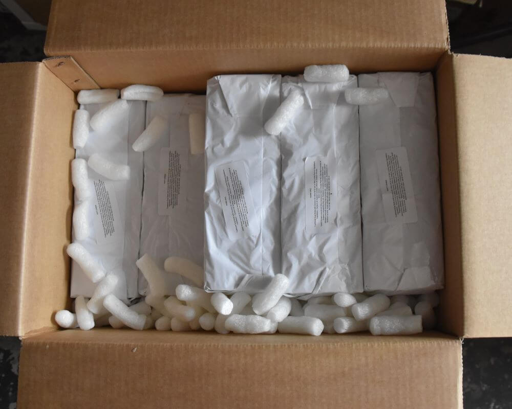 Image of an open box , seen from the top, packed with soap bars wrapped in white paper, with packing peanuts on one side nd a few on top