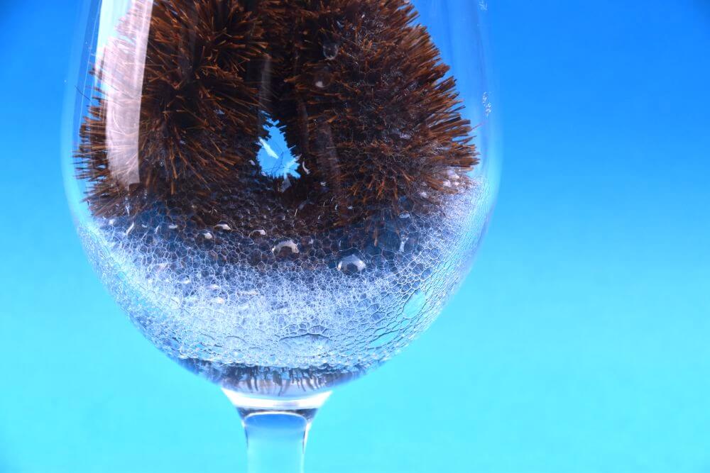 Image of a coconut washing up brush in a glass with foam at the bottom, against a blue background.