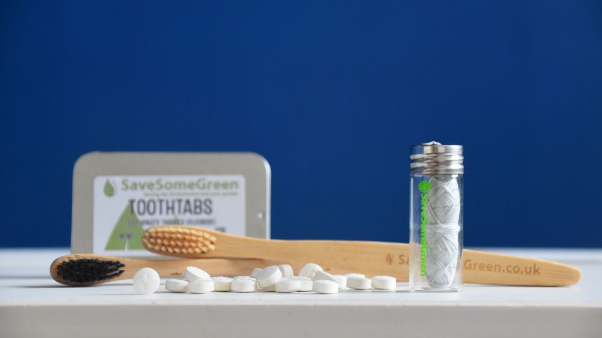 Image of a bottle of floss, some scattered toothtabs, two bamboo toothbrushes and a tin of toothtabs blurred in the background, on a white surface, against a blue background.