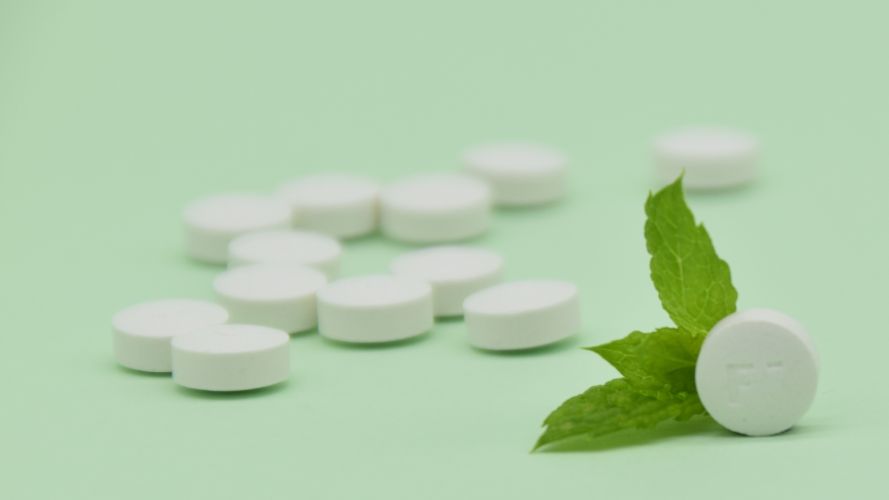 Image of tooth tabs, plastic free tooth paste alternative, some scattered around, one on the right hand side standing on its side together with some mint leaves.